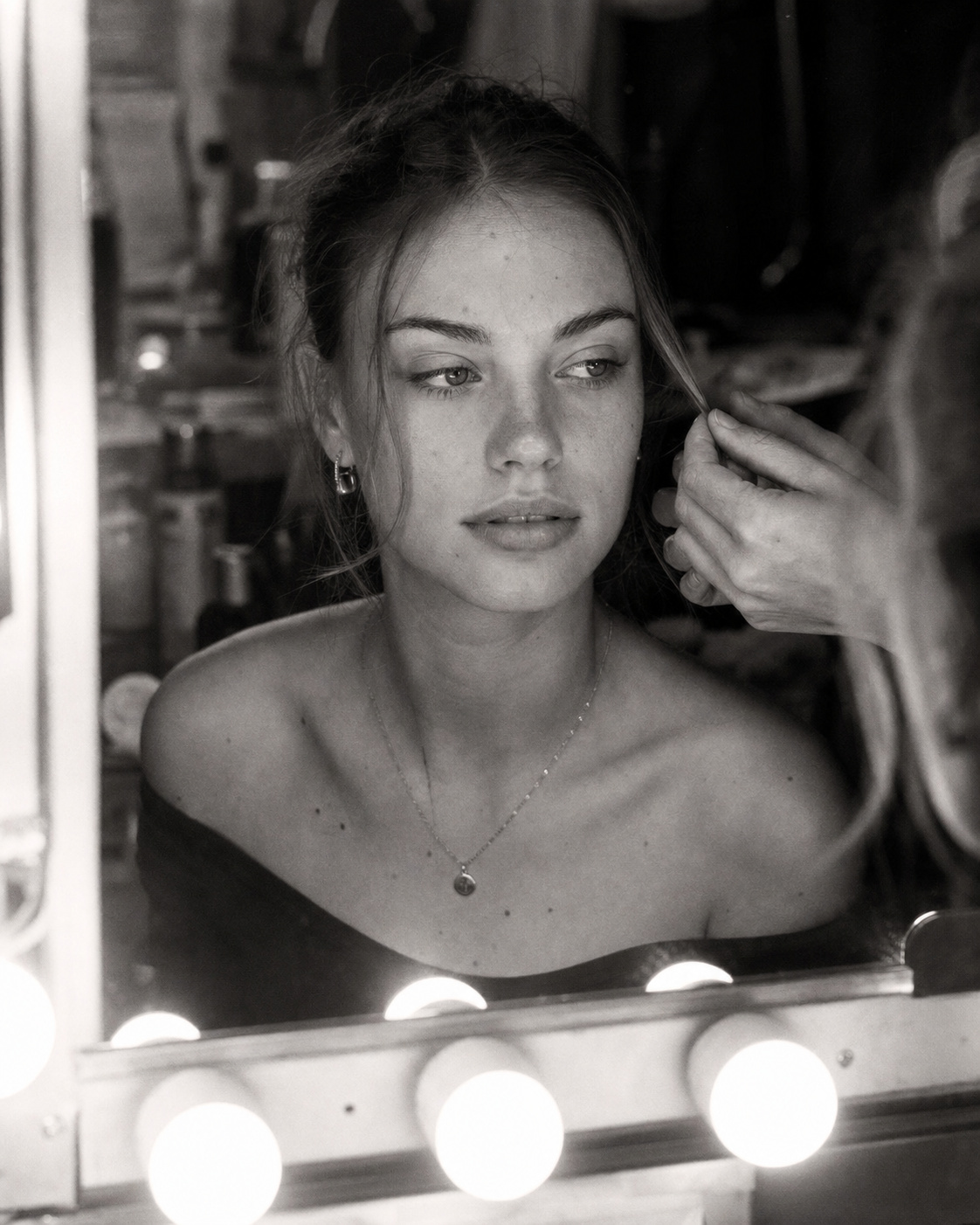 Black-and-white photo of a stylist smoothing a model&rsquo;s hair in a lit vanity mirror backstage