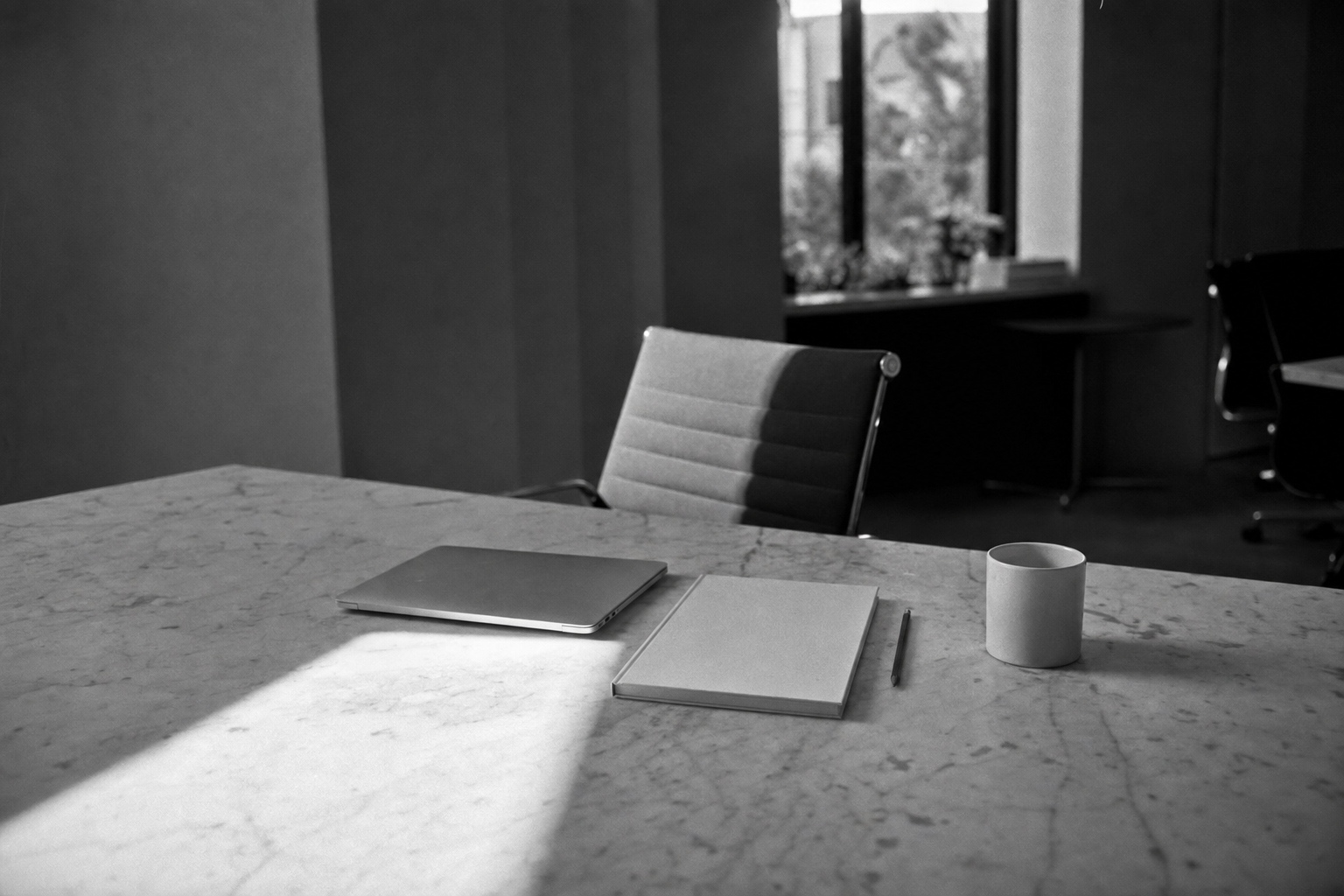 Black-and-white photo of an empty desk with a closed laptop and notebook at the end of a marble table in hard afternoon light