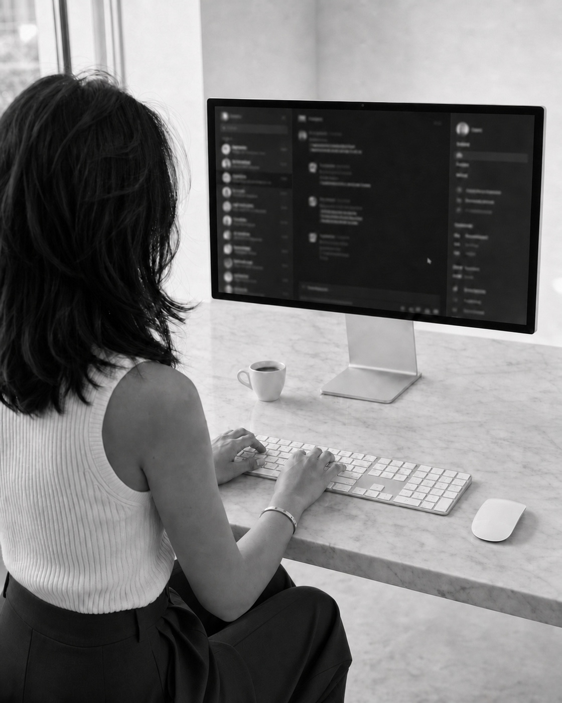 Black-and-white photo over a woman&rsquo;s shoulder as she types at a marble desk, message threads out of focus on the monitor