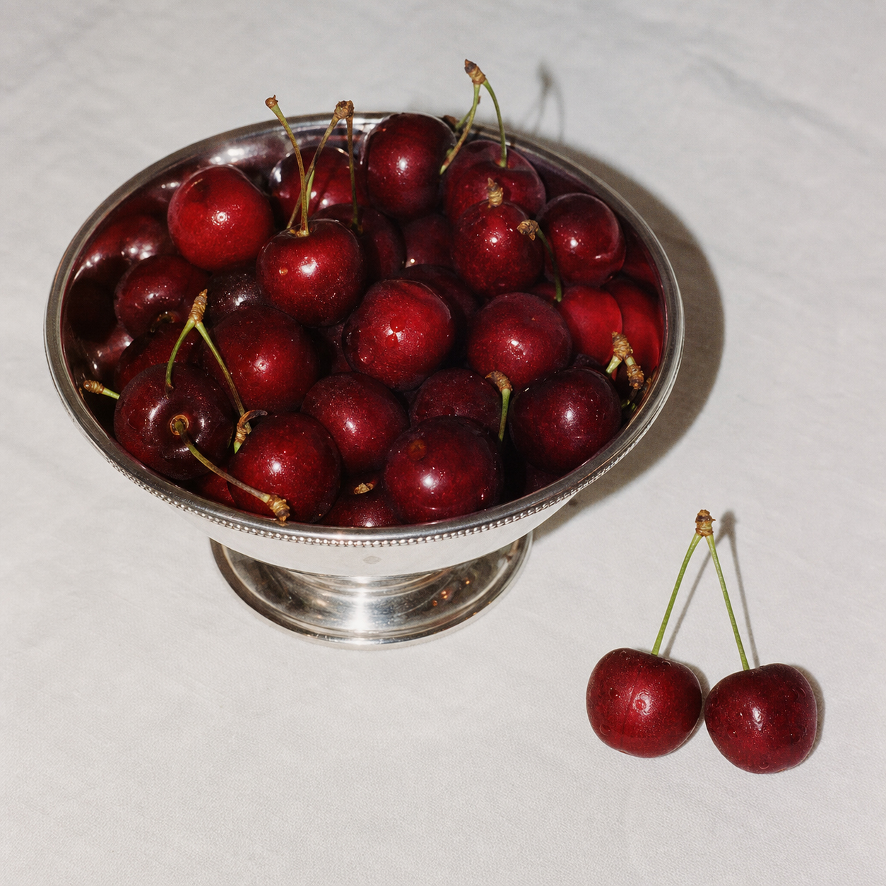 Fresh red cherries in a silver bowl on a white tablecloth with one pair beside it under hard flash