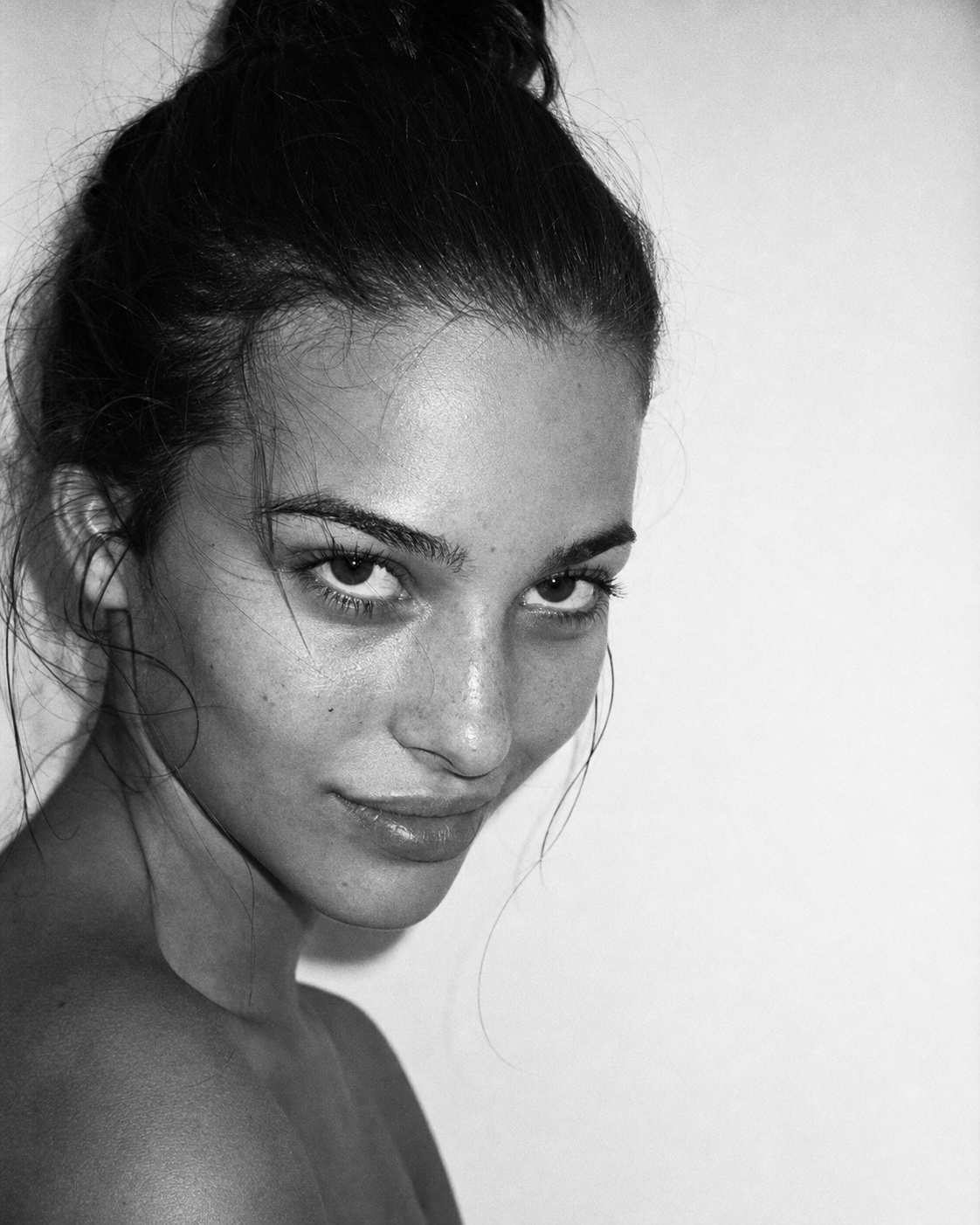 Black-and-white flash portrait of a model with minimal makeup smirking against a white wall