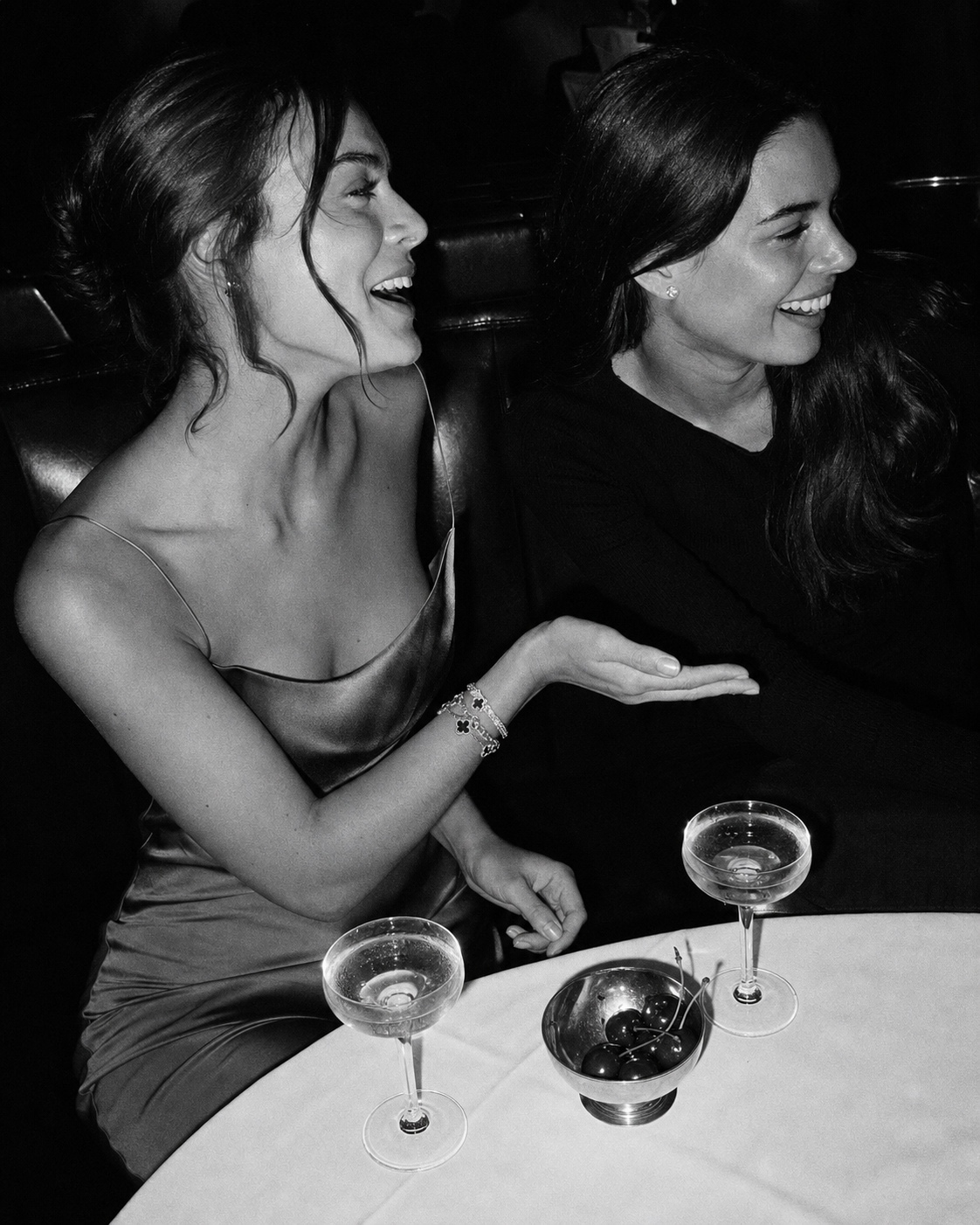 Black-and-white photo of two young women laughing together on a restaurant banquette with champagne coupes and a silver bowl of cherries